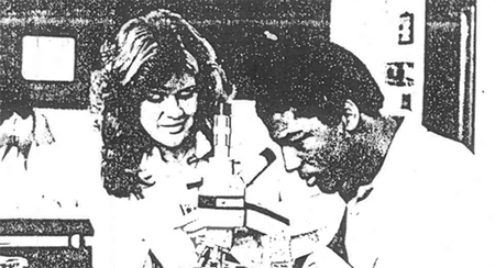 An old black and white image showing two people looking at a microscope. One is looking through the lens at a slide underneath while the other looks on.