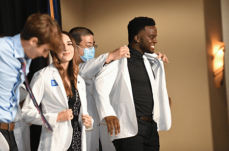 A student stands, smiling proudly, as he is fitted with his new white coat during the School of Medicine's 2022 ceremony.