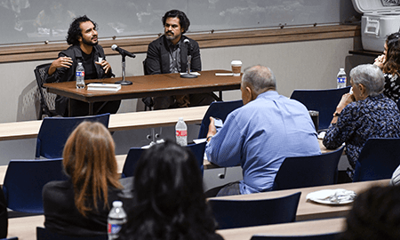 Author, poet and activist Javier Zamora sits at a table in front of an auditorium while discussing his memoir, Solito. Zamora, wearing a dark suit and sporting a trim beard, is gesturing with his hands as the audience appears abosorbed in his story.