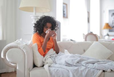 A woman sits on her couch sneezing and covered in a blanket