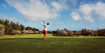 Photo of a man golfing, taken from behind as he swings with the blue sky in the background.