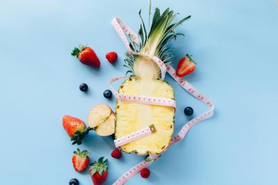 Photo of a pineapple sliced in half surrounded by berries all wrapped in a measuring tape to symbolize healthy eating and weight loss. 