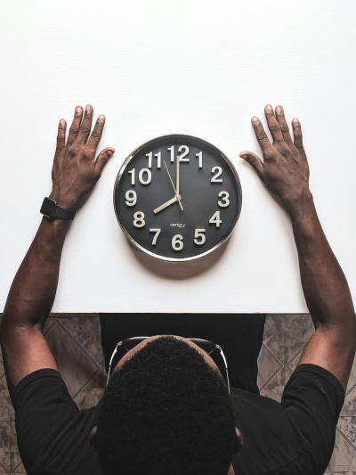 Photo taken from above of a clock face up on a table with a man's hands resting on either side of the clock. 
