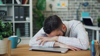 Man sleeping on top of closed laptop at desk.