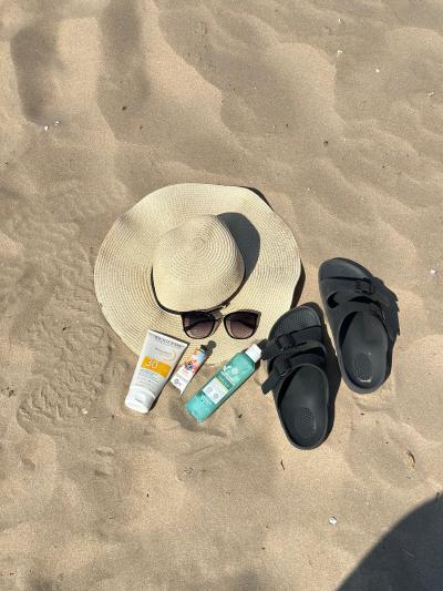 Close up shot of a sun hat, sunglasses, sandals and types of sunscreen on the sand at the beach.