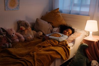Young child sleeping in her bed with lamp on. Surrounded by stuffed animals.