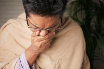 Photo of a man coughing, covering his mouth. He is wrapped in a blanket to indicate he isn't feeling well. 