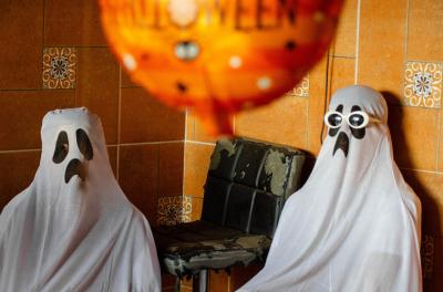 Two people wearing white sheet ghost costumes sitting in a corner of a room. 
