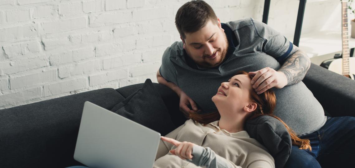 Overweight couple sitting a couch and looking at a laptop computer.