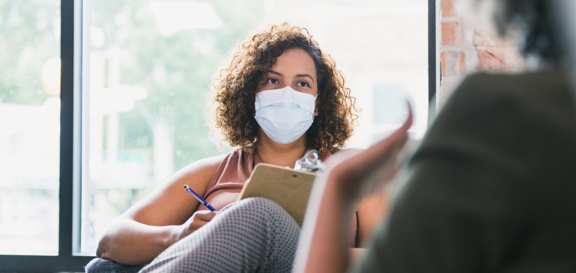 Masked woman holding a pen and clipboard having a conversation with a person not seen.