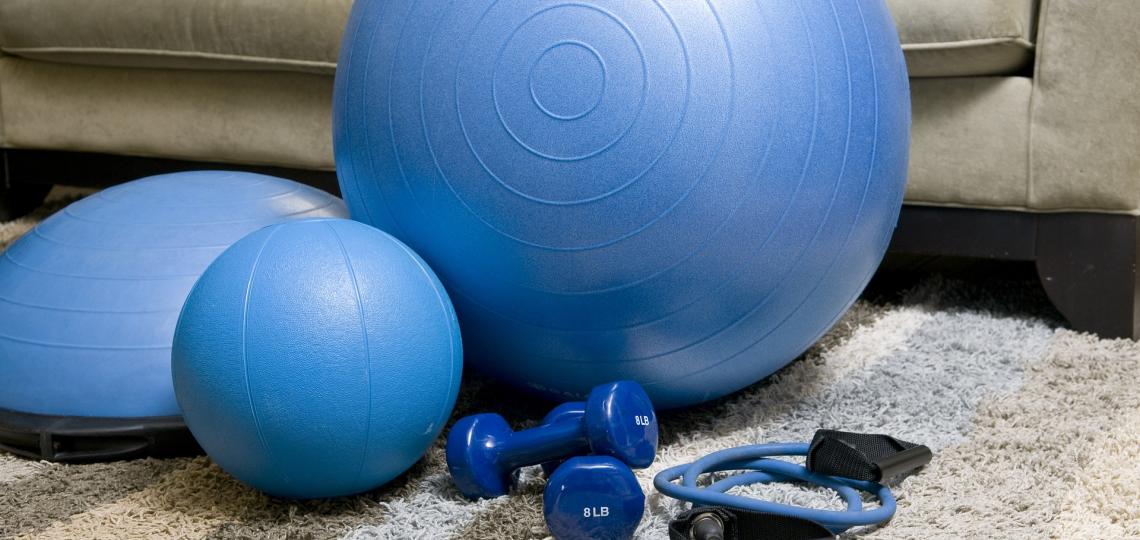 Blue exercise ball, weights and band sitting on the floor in front of a grey couch. 
