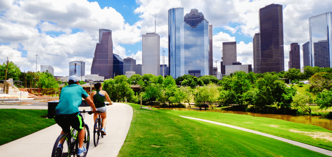 Two people ride bicycles toward downtown Houston, traveling across green grass with a blue, cloud-filled sky
