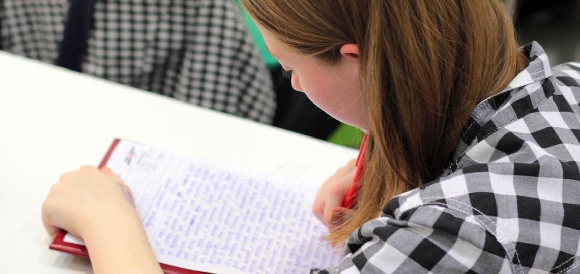 A photo of a girl, over he shoulder, looking down on a sheet of paper she is writing on. 
