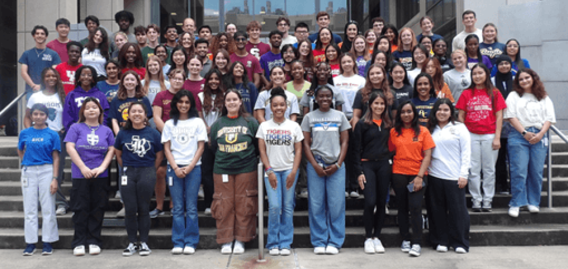 A large group of teenage students posed in rows on stairs, smiling. Many are wearing shirts with college logos.