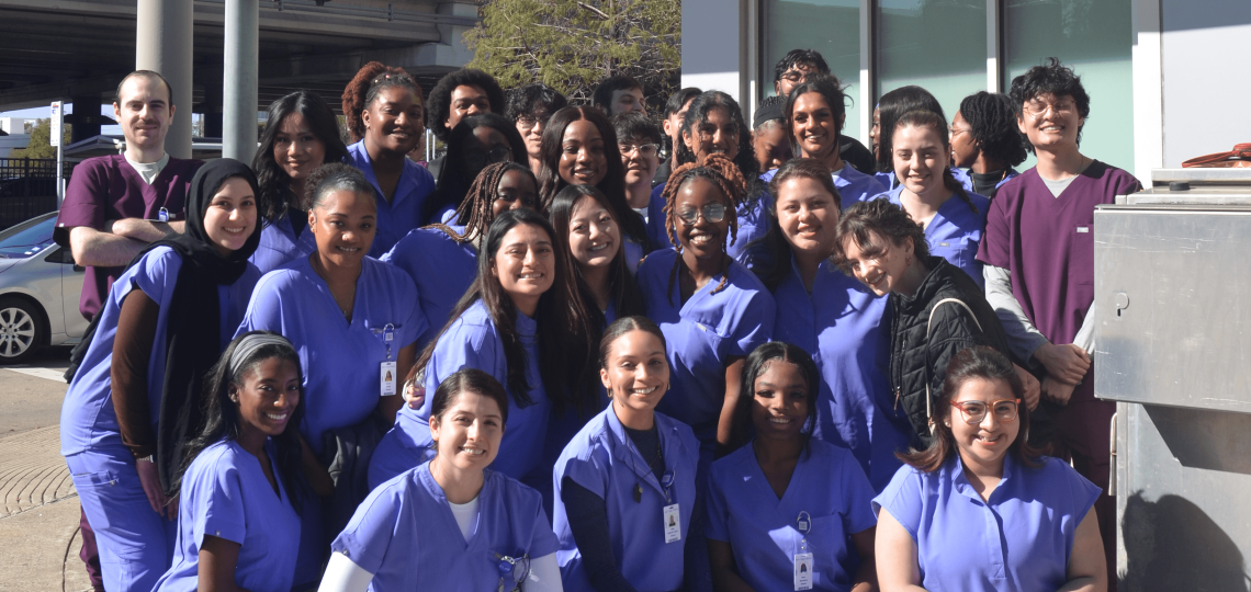 A group of students in blue or purple scrubs smiling outside a clinic.