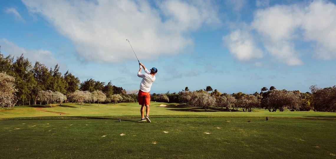 Photo of a man golfing, taken from behind as he swings with the blue sky in the background.