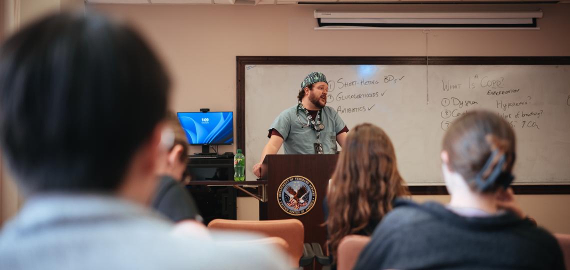 man in scrubs speaking at a podium in a classroom of people