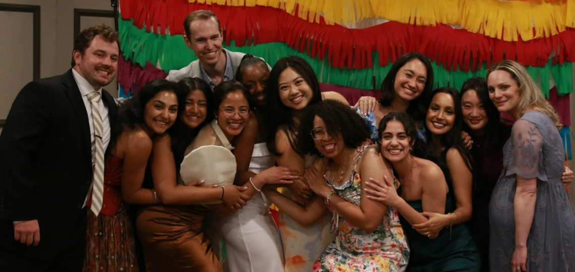 A group of people huddled together and hugging while facing the camera. There's a multi-colored banner in the background.