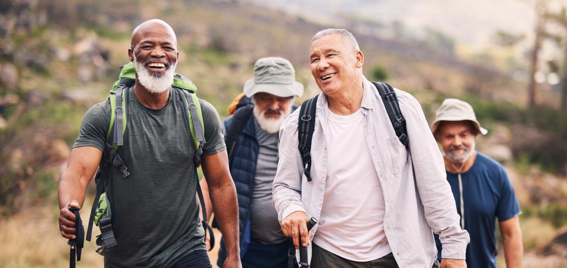 A group of older men hiking together on a scenic trail, smiling and enjoying each other's company. They are dressed in casual outdoor gear with backpacks and walking sticks, surrounded by nature and mountainous terrain.