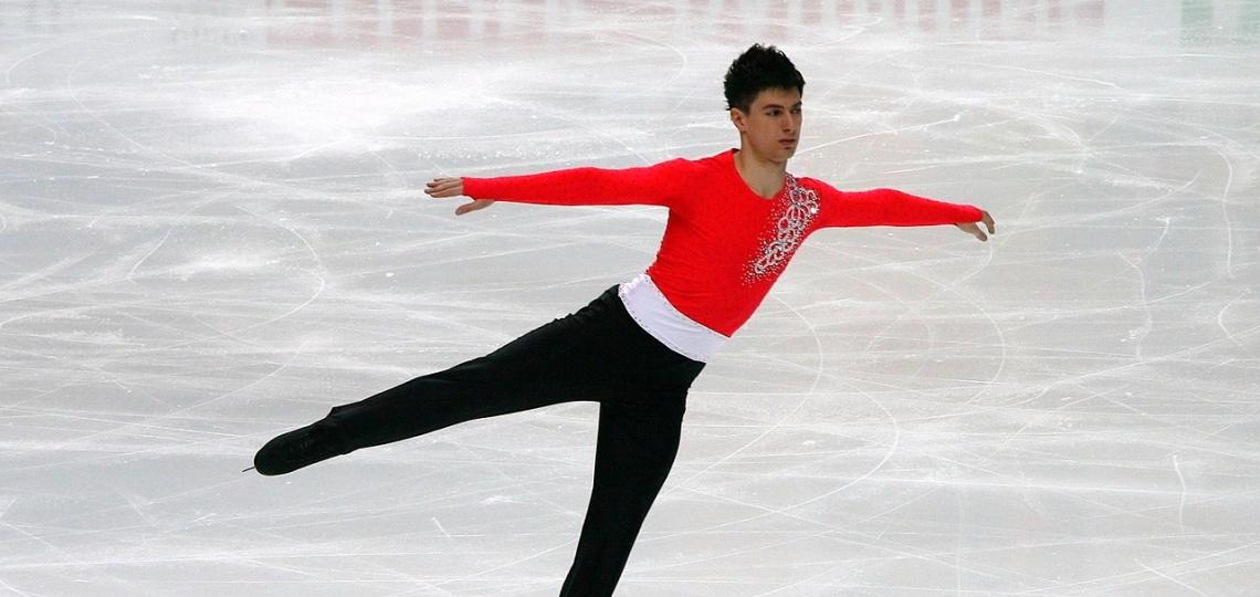 A man in black pants and a red top figure skating with arms outstretched and one leg in the air.
