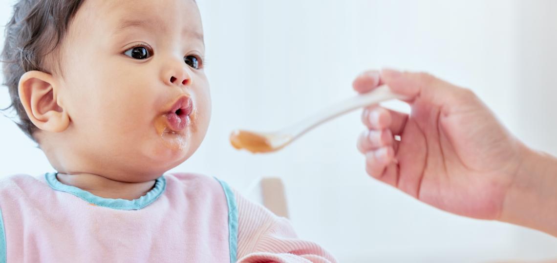 Baby eating solid baby food from a spoon being held by caretaker.