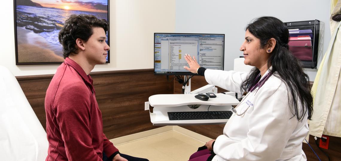 A doctor consults with a young man in a medical exam room, pointing at information on a computer screen.