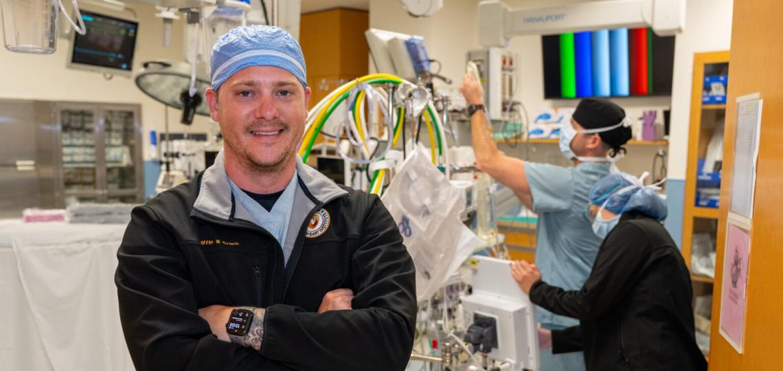 A smiling student with arms crossed standing amongst a cardiovascular perfusion machine being examined by two other students