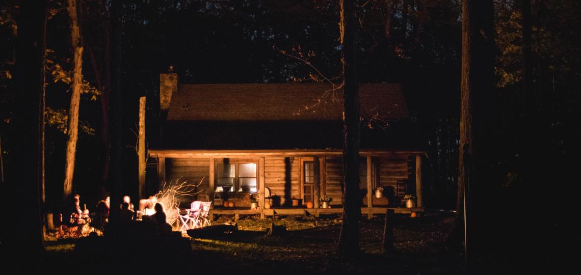The image is a photo taken at night of a cabin in the woods, a group of people are sitting around a camp fire in front of the cabin.