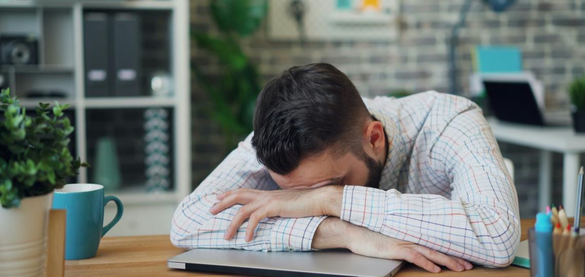 Man sleeping on top of closed laptop at desk.