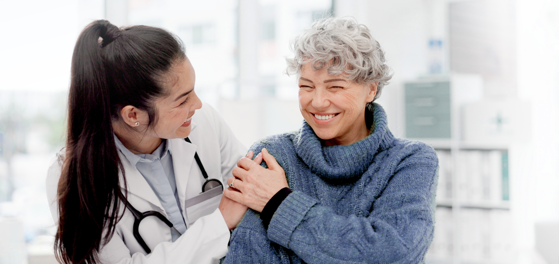A smiling healthcare professional in a white coat and stethoscope gently holds the arm of an older woman in a blue sweater, who is smiling warmly in a bright clinical setting.