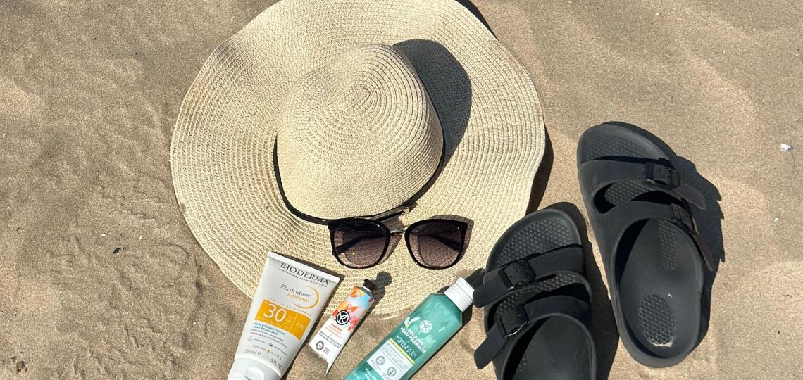 Close up shot of a sun hat, sunglasses, sandals and types of sunscreen on the sand at the beach.