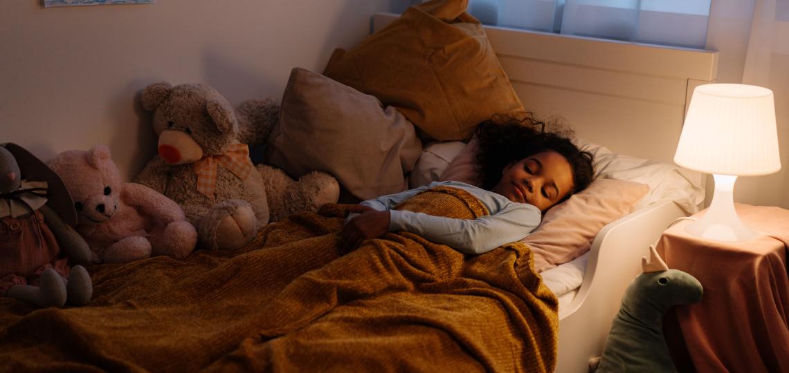 Young child sleeping in her bed with lamp on. Surrounded by stuffed animals.