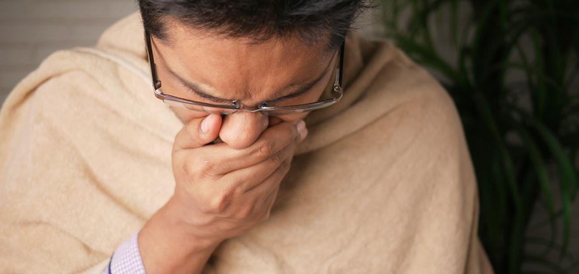 Photo of a man coughing, covering his mouth. He is wrapped in a blanket to indicate he isn't feeling well. 