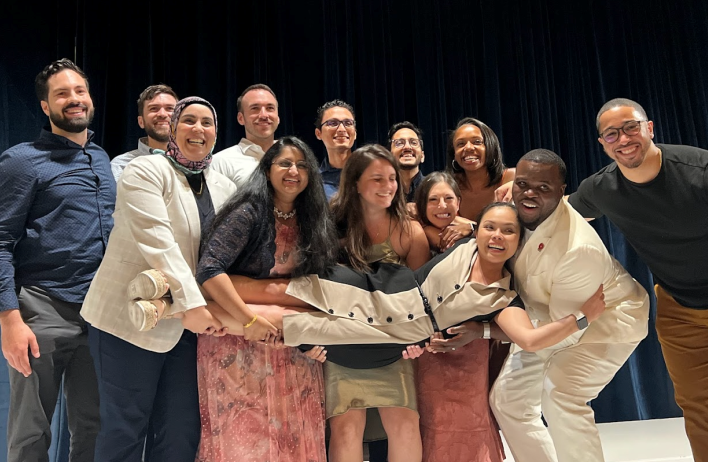 Emergency Medicine Residency Wellness members gather during a group meeting. They stand together in a row, with the group holding one person aloft in a horiztonal reclining position.