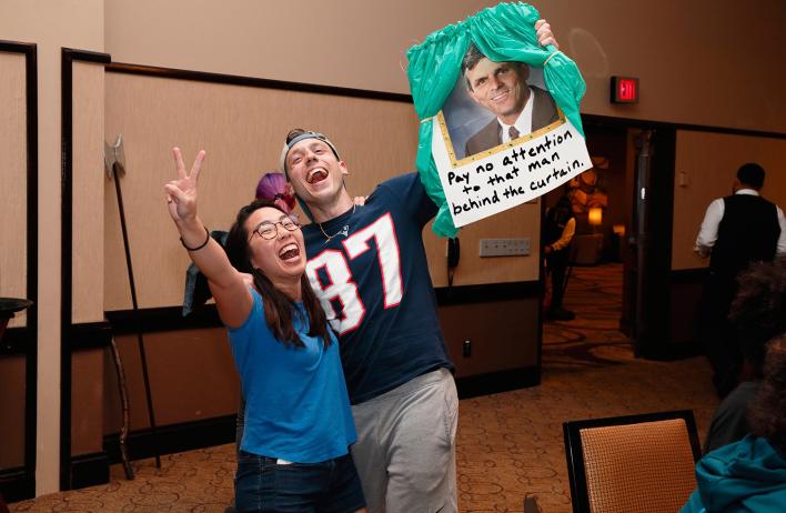 Two people cheering. One flashes a peace sign, the other holds a photo of a man with the words "Pay no attention to that man behind the curtain."