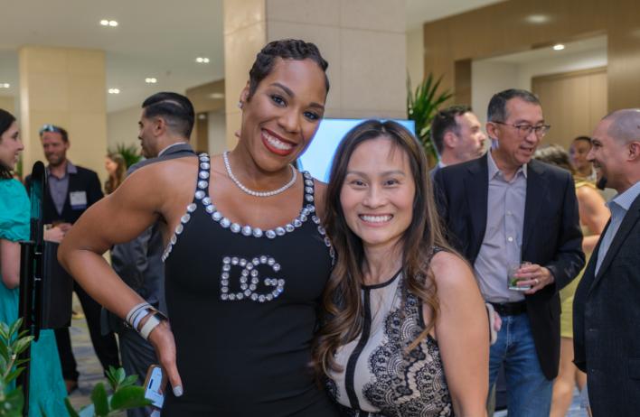 Two people smiling for the camera with a large group of laughing people behind them