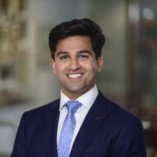man with dark hair in a blue suit and tie smiling