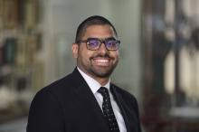 Male with short dark hair hair and glasses smiling. Wearing a black suit jacket with black tie and white button up shirts, blurred background 