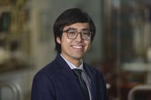 Male with should length dark hair and glasses smiling. Wearing a navy blue suit jacket and tie, blurred background