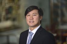 Male smiling with short dark hair, wearing a black suit jacket and silver tie with a white button up shirt, blurred background
