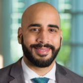 Male, bald, smiling, with facial hair. Wearing a dark gray suit jacket