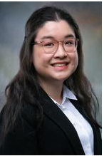 Female with long dark hair and glasses, smiling. Wearing dark suit jacket and white button-down shirt.