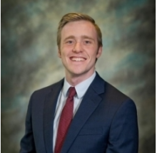 Male with blonde hair smiling. Wearing a red tie and dark blue suit jacket