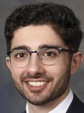 man with dark hair and facial scruff smiling