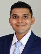 man with dark hair in a blue suit and light blue tie smiling
