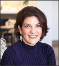 Portrait of a person with short dark hair, wearing a dark blue textured top and large earrings, in an indoor setting