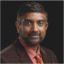 man in a brown blazer with dark hair smiling lightly