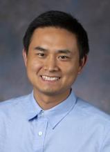 man with dark hair in a light blue button down, smiling
