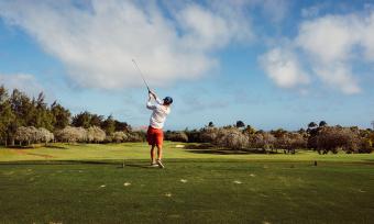 Photo of a man golfing, taken from behind as he swings with the blue sky in the background.