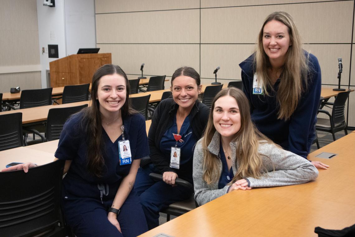 Four people seated in a classroom smiling while wearing dark blue scrubs.